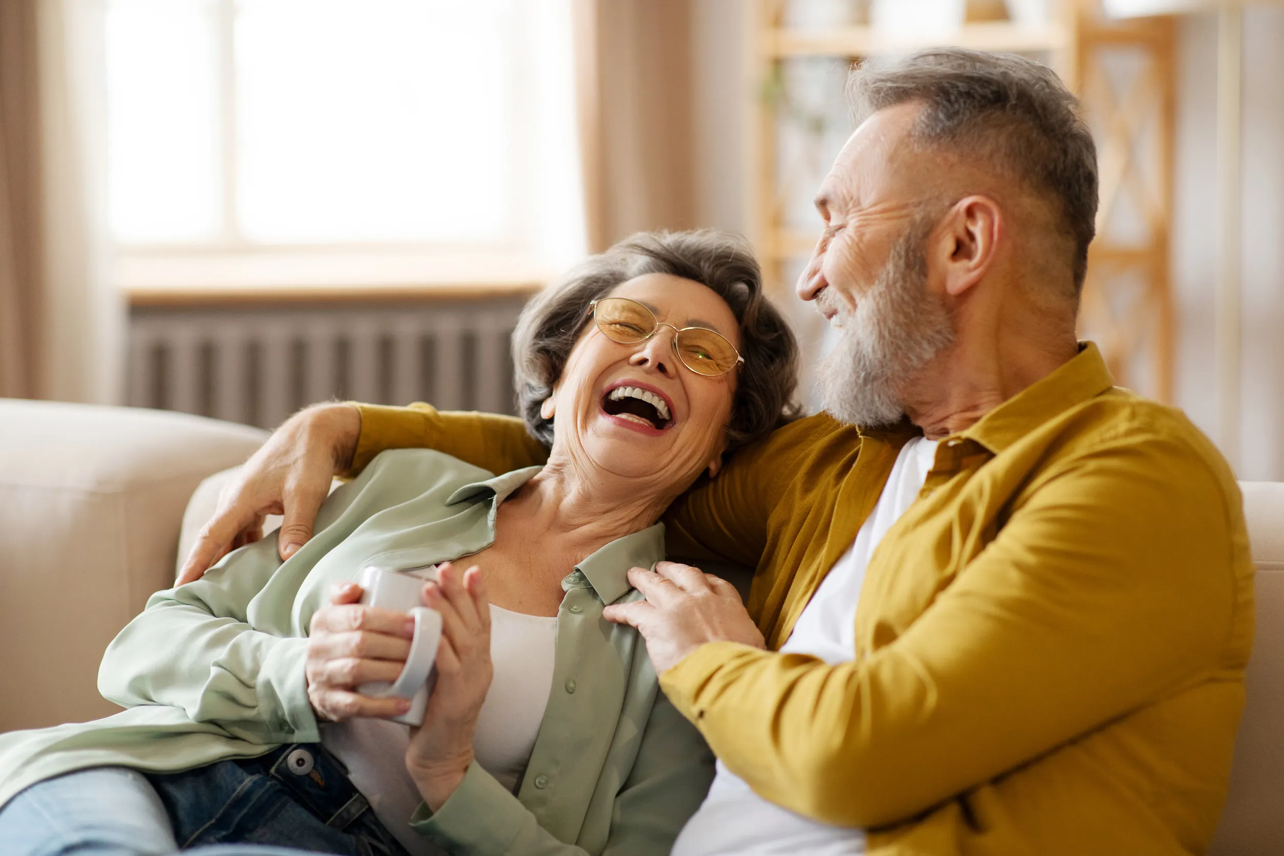 Senior spouses talking, sitting on sofa, woman holding cup of coffee and laughing to husband