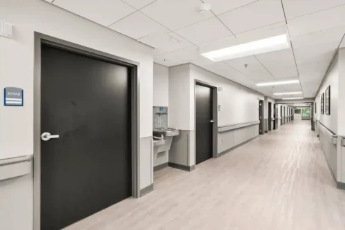 Long hospital corridor with beige flooring, white walls, and multiple black doors. Bright overhead lighting; a calm, sterile atmosphere prevails.