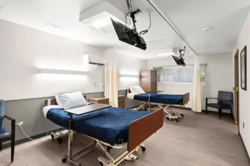 A hospital room with two beds, each with blue blankets and white pillows, separated by a curtain. Wall-mounted TVs face each bed, and a window lets in daylight.