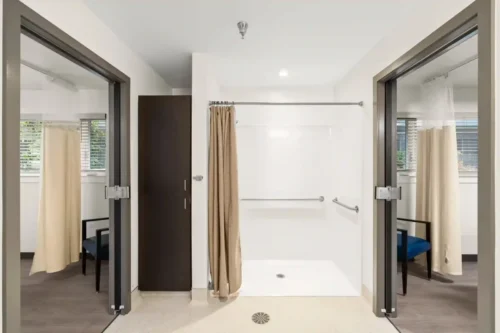 Modern accessible bathroom with a central walk-in shower, beige curtain, and grab bars. Flanked by two rooms with chairs, creating a functional, inviting space.