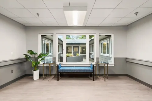Modern, bright waiting area with a blue bench between two gold tables. Large window reveals greenery outside, adding a tranquil atmosphere.