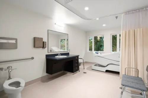A bright, accessible bathroom with a toilet, sink, mirror, and a high-sided bathtub. Safety bars and a shower chair suggest a supportive and calming environment.