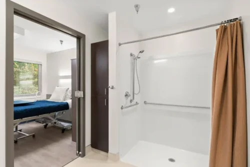 Accessible bathroom with a walk-in shower featuring grab bars and a brown curtain. Adjacent room holds a medical bed with a blue cover, creating a clinical, calm setting.