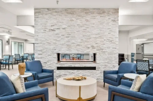 Contemporary lounge with blue armchairs, a round white coffee table, and a central stone fireplace. Bright and inviting atmosphere with neutral tones.