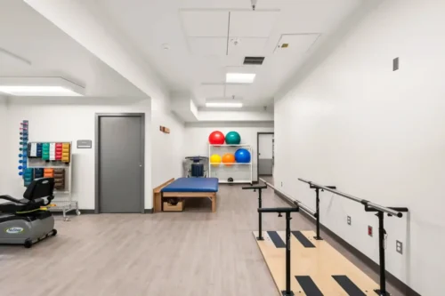 Bright physical therapy room with exercise bike, padded table, vibrant balance balls on shelves, parallel bars, and neutral walls, conveying a clinical, organized setting.